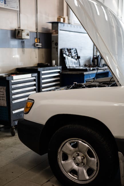 White car with hood open in a professional auto repair shop. Toolbox and equipment visible.